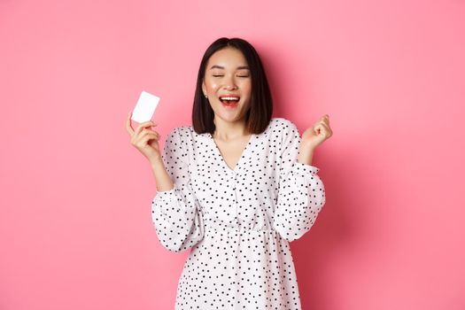 Shopping concept. Excited asian woman feeling satisfaction, holding credit card and rejoicing, standing over pink background.
