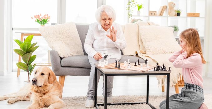 Little beautiful girl and her grandmother smiling during chess game in the light room with golden retriever dog on the floor