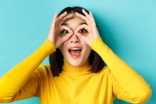 Close up of funny asian woman looking through hand binoculars with surprised face, see something amazing, standing over blue background.