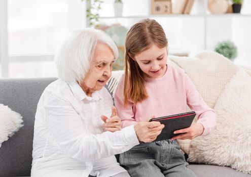 Grandmother and granddaughter sitting on the sofa and looking at the tablet together