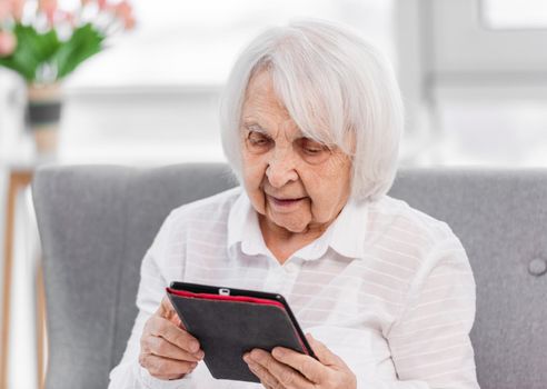 Elderly woman sitting on the sofa at home and reading from the tablet screen