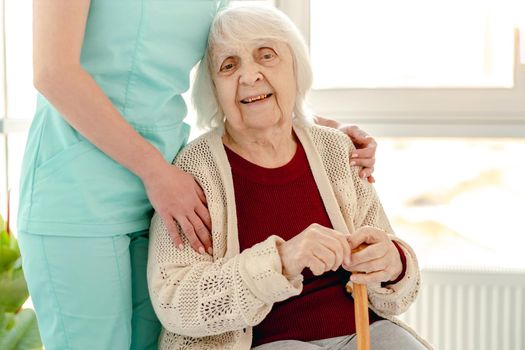 Happy old lady sitting on chair next to cuddling nurse in home for elderly