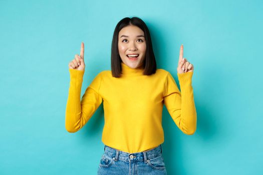 Shopping concept. Beautiful asian woman in yellow sweater pointing fingers up at logo, looking at promotion with happy smile, standing over blue background.