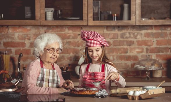 Granny helps granddaughter sprinkling sugar powder with sieve on homemade cake