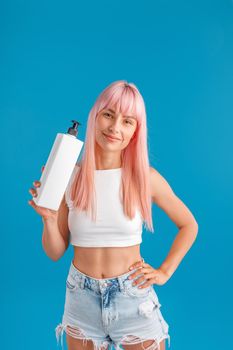 Beautiful woman with smooth pink hair smiling at camera and showing pump bottle with hair mask or shampoo, standing isolated over blue studio background. Beauty and hair care concept