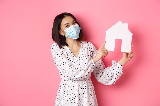 Covid-19, real estate and lifestyle concept. Cute asian woman in face mask selling houses, showing model of home and looking at camera, standing over pink background.