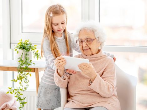 Cute school girl helping old lady with gadget in light room