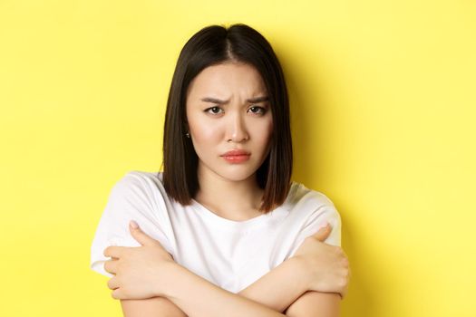 Sad timid girl comforting herself, hugging body and frowning upset, standing again yellow background.