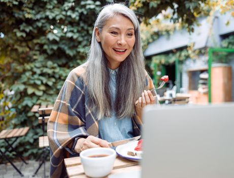 Happy grey haired senior Asian lady eats toast with fresh strawberries watching video via laptop at table on outdoors cafe terrace on autumn day