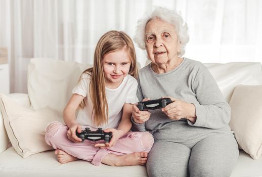 Smiling grandmother with little granddaughter playing games together with gamepads