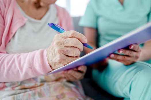 Elderly woman and nurse hands sign documents at home closeup. Healthcare worker girl and senior female person with papers indoors