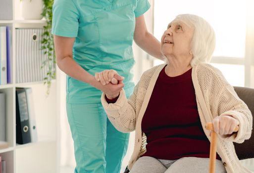 Old lady leaning on nurse hand to stand from chair in nursing home