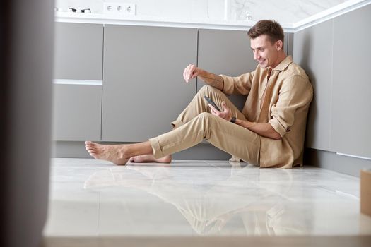 Beautiful caucasian man is sitting on the floor and using smartphone on light modern kitchen