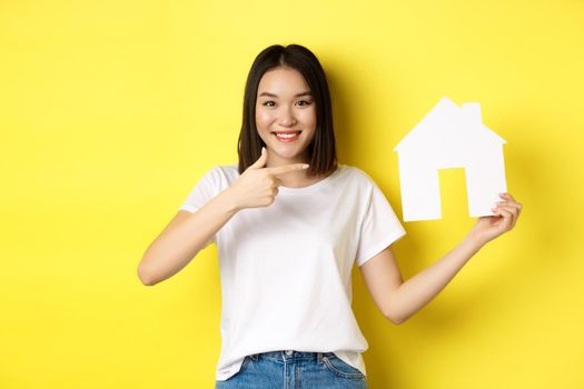 Real estate and insurance concept. Cheerful asian woman smiling, pointing at paper house cutout, recommend agency logo, standing over yellow background.