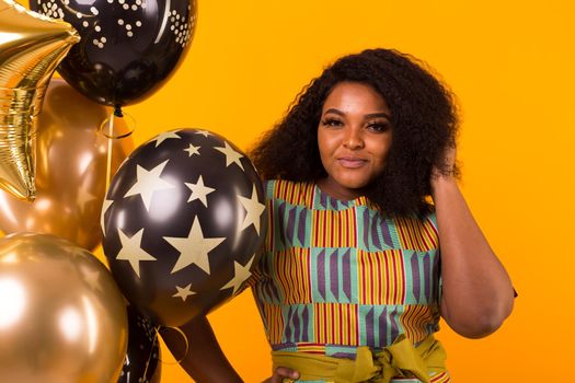 Portrait of smiling young African-American adult woman looking sweet on yellow background holding balloons.