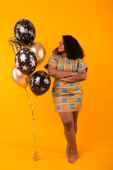 Portrait of smiling young African-American adult woman looking sweet on yellow background holding balloons.