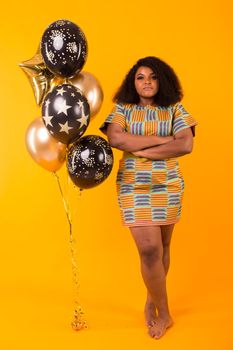 Portrait of smiling young African-American adult woman looking sweet on yellow background holding balloons.