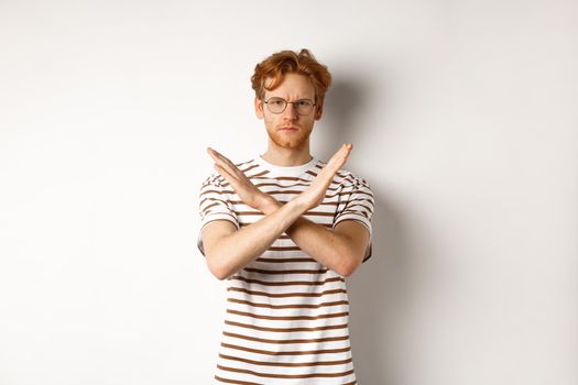 Serious young man with ginger hair frowning, showing stop gesture, making cross to prohibit something bad, disagree with you, standing over white background.