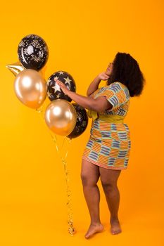 Portrait of smiling young African-American adult woman looking sweet on yellow background holding balloons.
