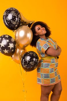 Portrait of smiling young African-American adult woman looking sweet on yellow background holding balloons.