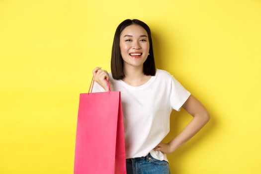Beautiful asian female model in white t-shirt, holding shopping bag and smiling satisfied, standing over yellow background.