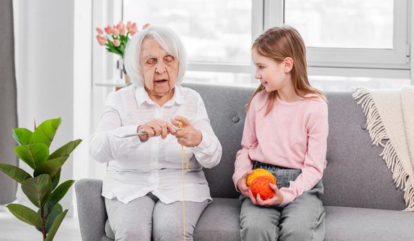 Grandmother holds needles and teach to knit preschooler granddaughter with thread ball in her hands