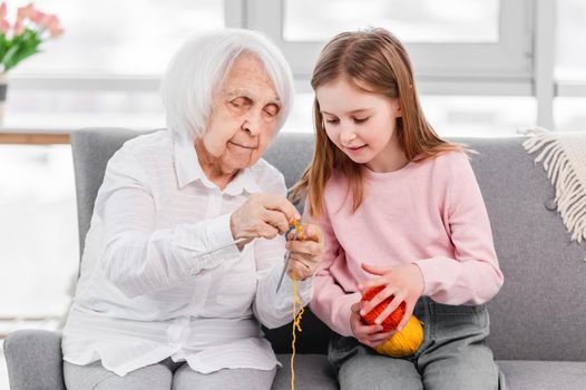 Grandmother taeching granddaughter knitting. Elderly woman sitting on the sofa with kid girl and holding wool threads