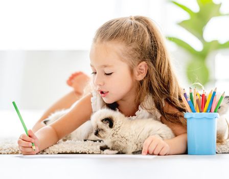 Child girl painting with ragdoll kittens and lying on the floor. Little female person drawing with colorful pencils and kitty pets close to her at home