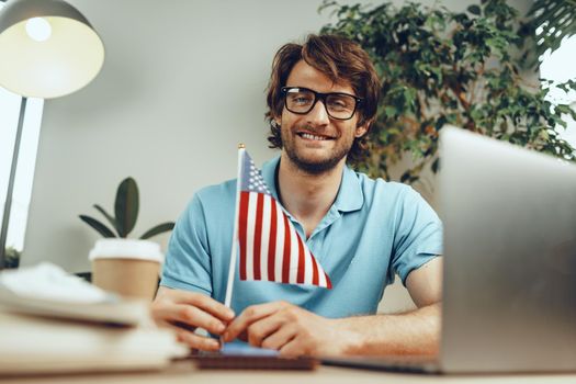 Young bearded businessman sitting at table with laptop and american flag, patriot man