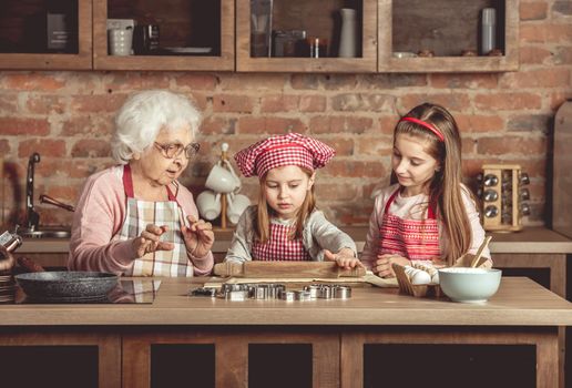 Grandma and granddaughters are spreading dough using a rolling pin