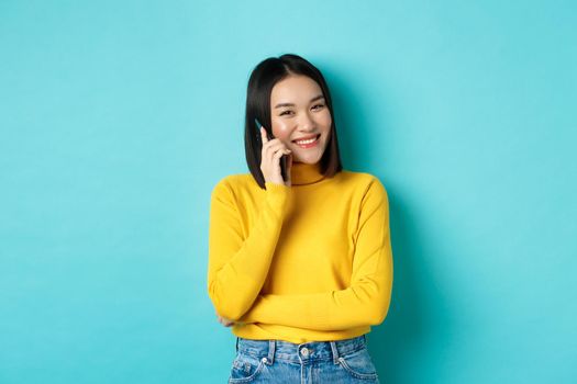 Young stylish asian woman talking on phone, calling friend and smiling, standing with smartphone against blue background.