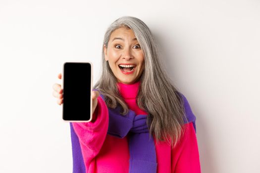 Online shopping. Close up of stylish asian senior woman extending hand with mobile phone, showing blank smartphone screen and smiling, standing over white background.