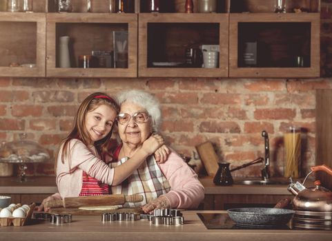 Little granddaughter hug her happy grandmother sitting at rustic kitchen at home