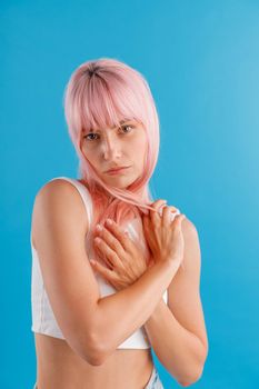 Beautiful young woman looking at camera, wrapping her pink hair around her neck while posing isolated over blue studio background. Beauty, hair care concept