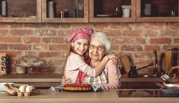 Little granddaughter hug her happy grandmother sitting at rustic kitchen at home
