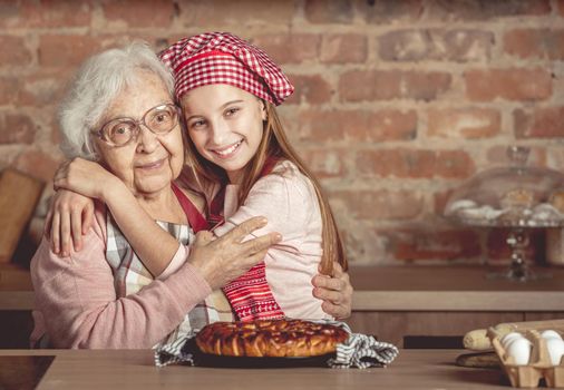 Little granddaughter hug her happy grandmother sitting at rustic kitchen at home