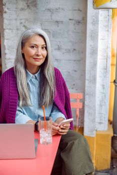 Positive mature Asian lady with straight hair holds mobile phone sitting at coral table with notebook on outdoors cafe terrace