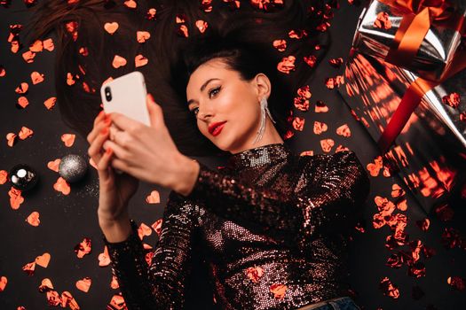 Top view of a girl lying in shiny clothes on the floor in confetti in the form of hearts and taking a selfie.