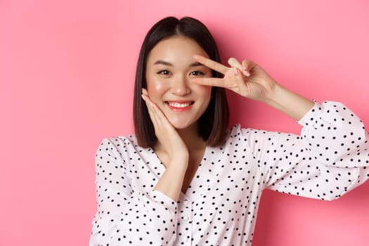 Beauty and lifestyle concept. Close-up of cute asian woman showing peace sign and touching cheek, smiling happy at camera, standing over pink background.