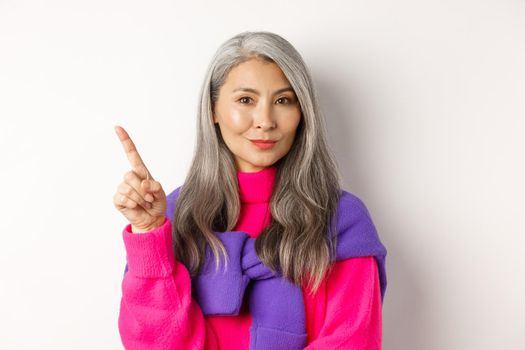 Close-up of asian beautiful mother with grey hair, smiling and pointing upper left corner, showing advertisement, standing over white background.