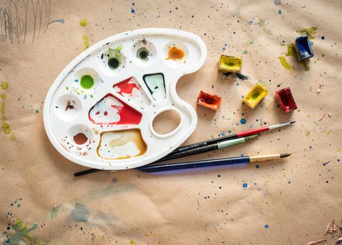 Plastic art palette with used brushes and opened paint boxes on paper-covered workshop table