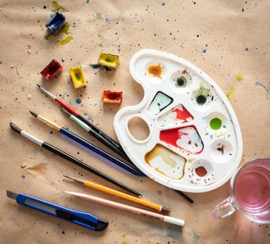 Plastic art palette with used brushes and opened paint boxes on paper-covered workshop table