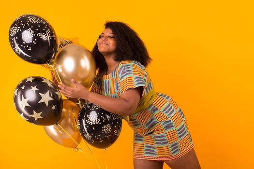 Portrait of smiling young African-American adult woman looking sweet on yellow background holding balloons.