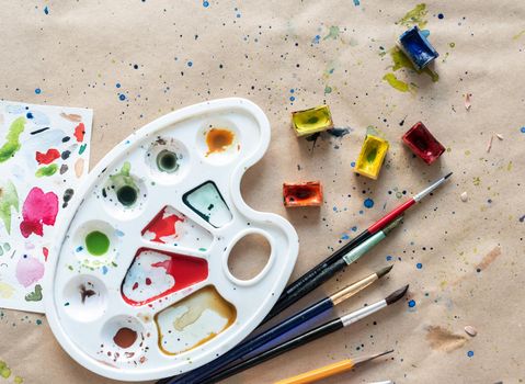 Plastic art palette with used brushes and opened paint boxes on paper-covered workshop table