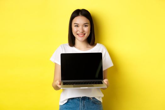 Young asian woman demonstrate online offer, showing laptop screen and smiling, standing over yellow background.
