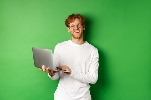 Cheerful redhead freelancer in glasses smiling at camera, working on laptop while standing over green background.