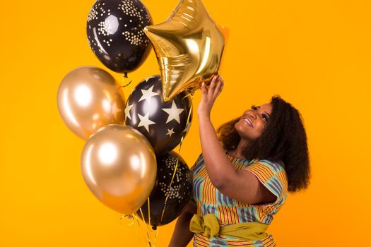 Portrait of smiling young African-American adult woman looking sweet on yellow background holding balloons.