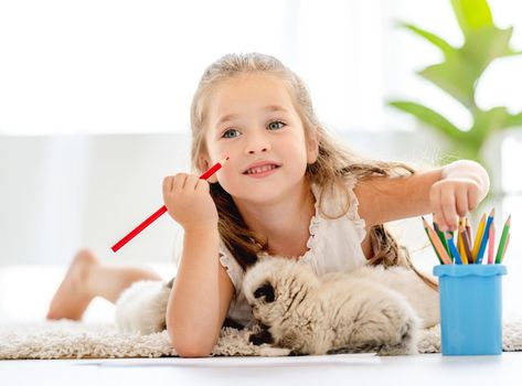 Child girl painting with ragdoll kittens on the floor and thinking. Little female person drawing with colorful pencils and kitty pets close to her at home
