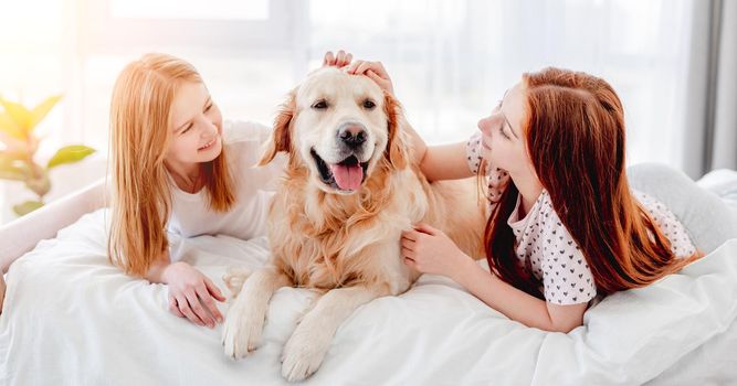 Two sisters lying in the bed and petting golden retriever dog in with sunlight in the room. Girls with pet staying at home. Beautiful portrait of friendship between human and animal