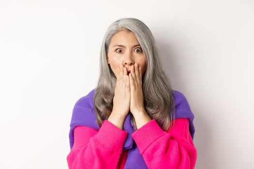 Close up of shocked asian old woman with grey hair, standing in stylish pink sweater, gasping startled and amazed, staring at camera, white background.